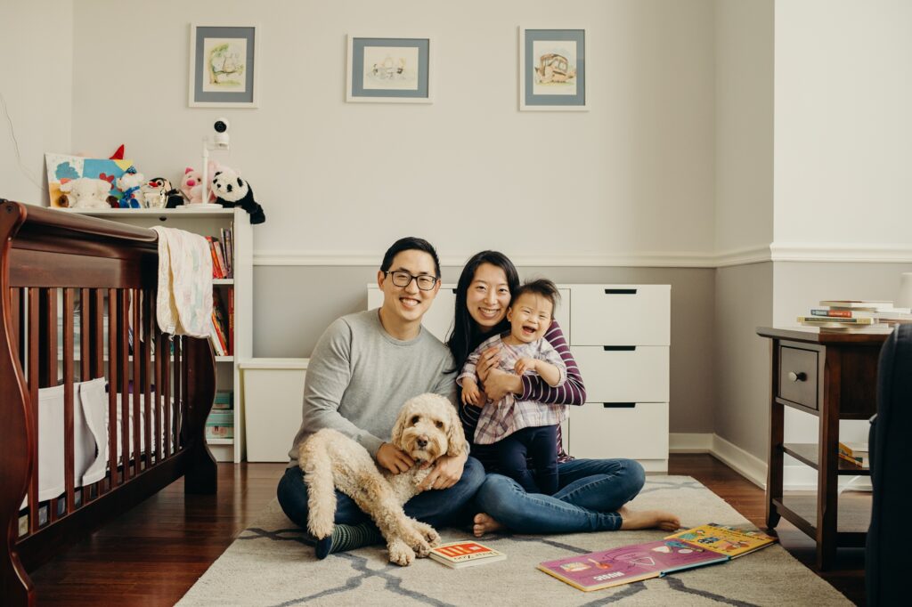 Family of three sits on the floor in a nursery with both baby and doodle dog in their laps. Everyone is smiling except harold the dog.