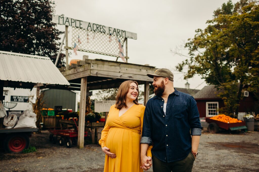 an expecting mother holding hands with her husband standing in front of a barn at Maple Acres Farm 