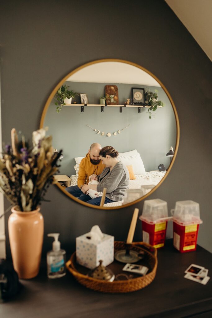 a mom and dad holding their newborn, photographed through a mirror hanging above their dresser. on their dresser is proof of their IVF treatment 
