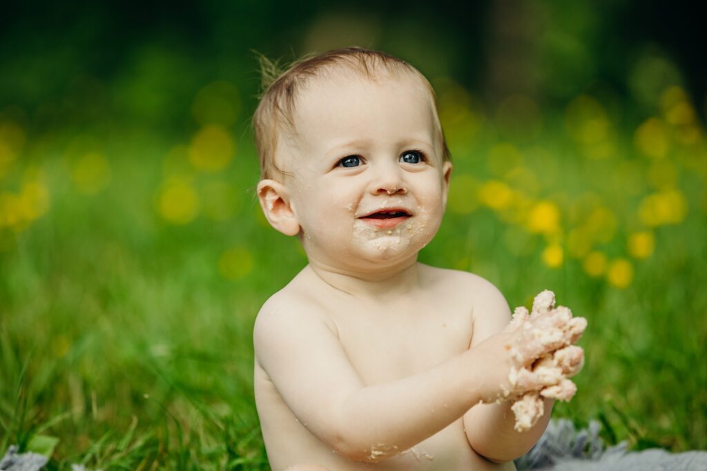 a close up photo of a baby boy, sitting in a field of yellow flowers (the flowers and background are blurred). He has cake crumbs and icing all over his mouth and hands.