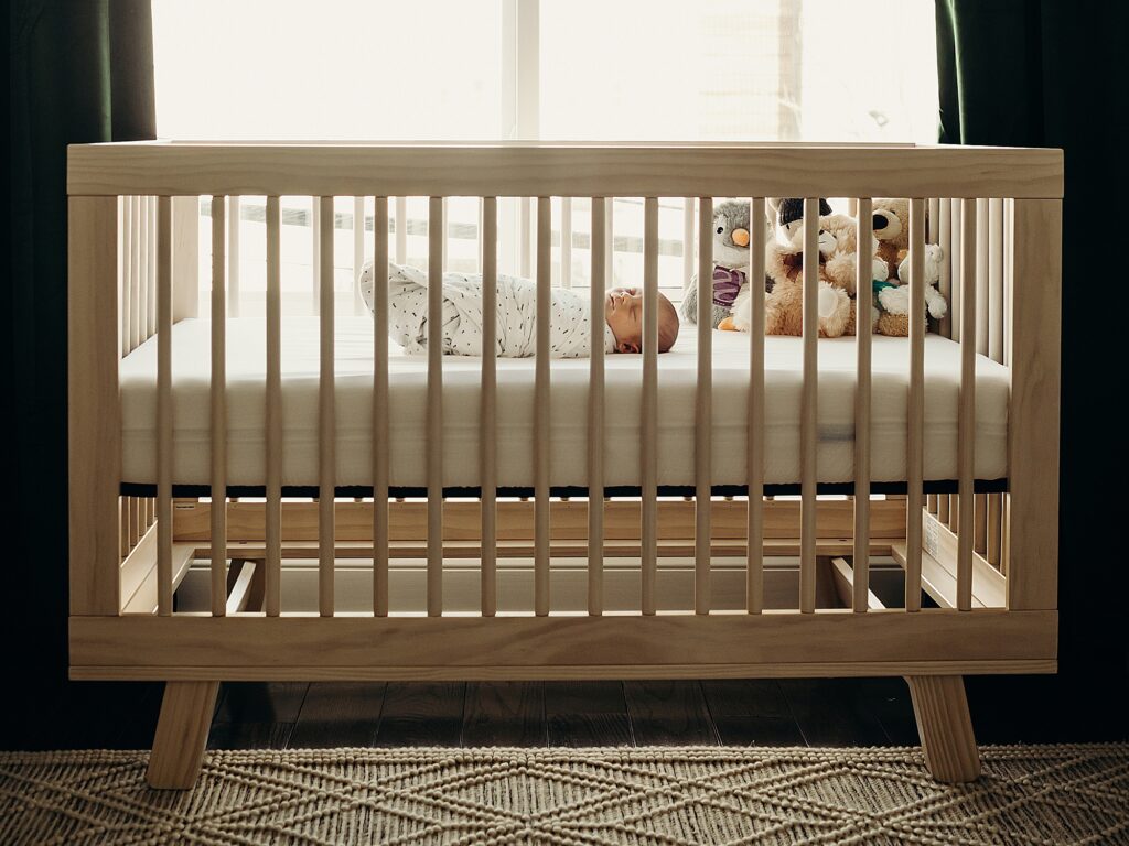 There is a swaddled baby asleep in his nice wooden crib, with a pile of stuffed animals in the corner of the crib. The crib is placed in front of a large window, as beautiful sunlights shines on the crib.