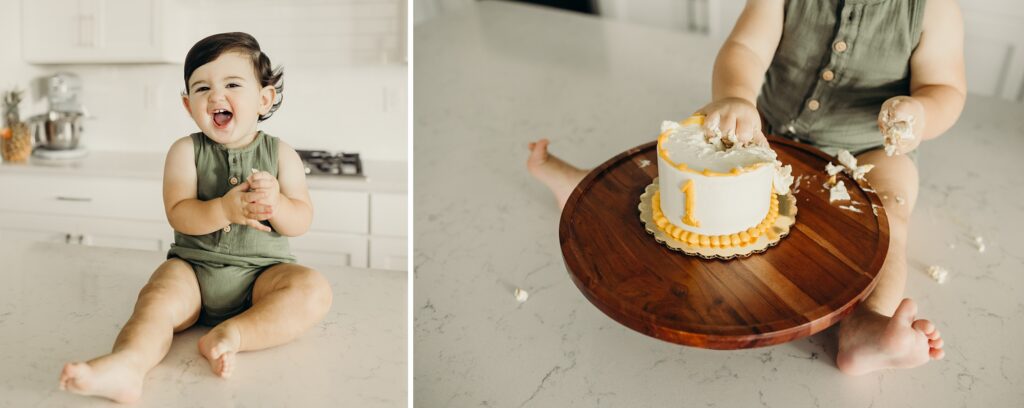 a collage of 2 photos. the photo on the left shows a baby boy sitting on a kitchen island, he is laughing and clapping his hands together as he looks into the camera. the right photo is just of the babys legs, as he still sits on the island. a cake stand is now between his legs, holding up a white and yellow cake that has the number 1 written on it. the baby boy's hand is messily grabbing the top of the cake. 