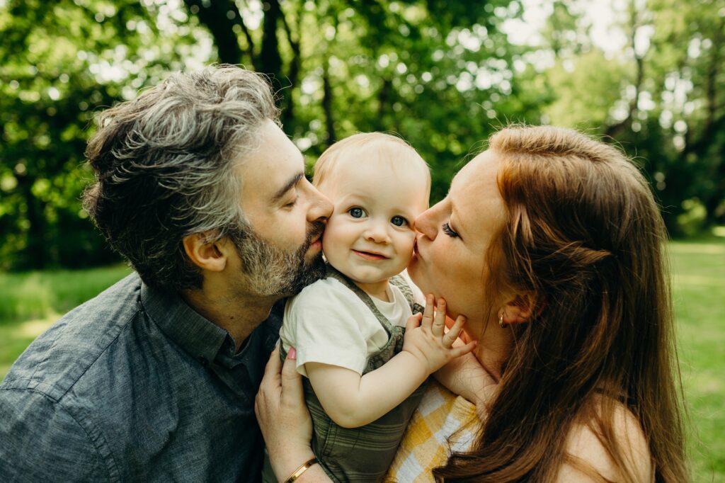a family outside. the dad and mother are on either side of their baby boy, kissing his cheeks. the baby looks into the camera smiling, as he holds his mothers face