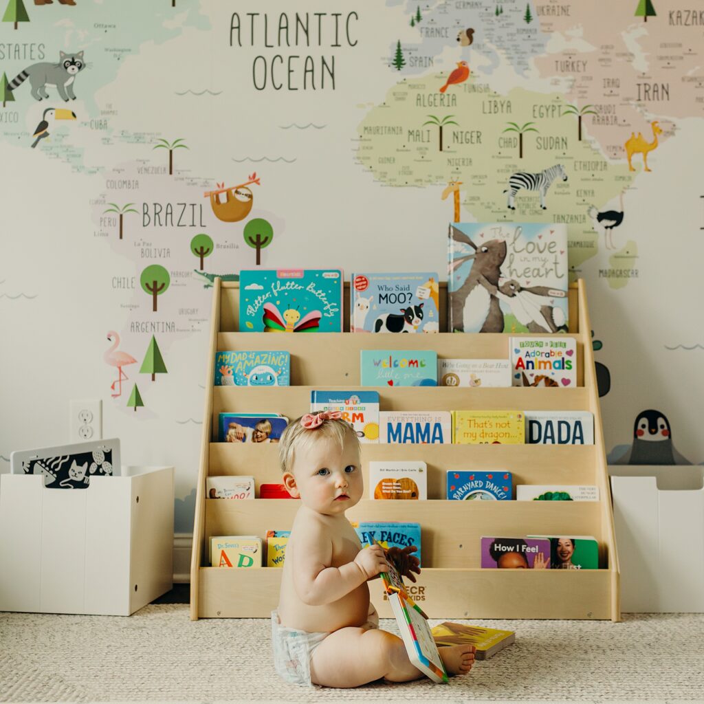 a baby girl in only a diaper is sitting in her playroom. the wallpaper is a colorful map of the world that has pictures of animals scattered across. There is a wooden layered bookshelf that holds numerous baby books. the baby is sitting in front of the book case, holding a book as she looks into the camera.