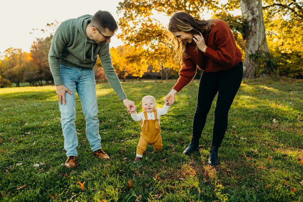 There is a family standing in a park during the autumn season, with beautiful fall foliage as their background. The father is standing on the left holding his 9 month old son's hand, while the mother is standing on the right, holding her sons other hand. The parents are looking down at their baby smiling, while the son looks into the camera smiling and taking a step forward. 