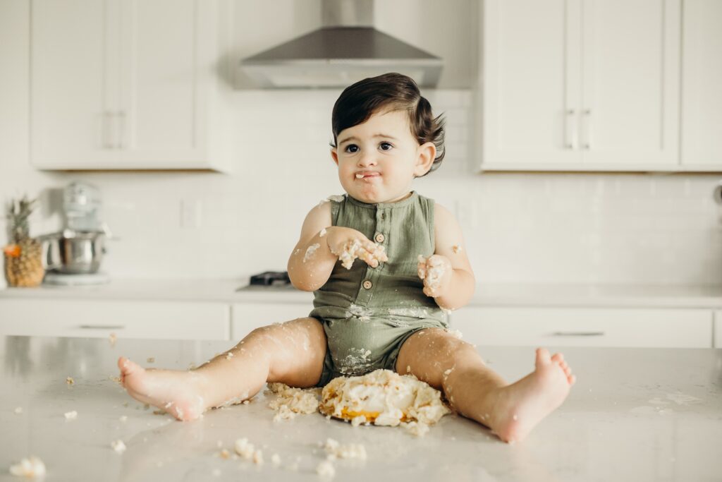 a baby boy is sitting on top of a white kitchen island. there is a crumbled up cake lying between his legs, as there are crumbs all over him and the counter. he is looking off to the left, sticking his tongue out to taste the crumbs on his face.