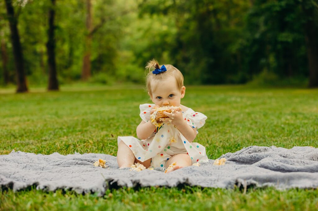 a baby girl sitting on a blue blanket in grass. she is staring directly into the camera and she is stuffing her face with a huge chunk of cake. 