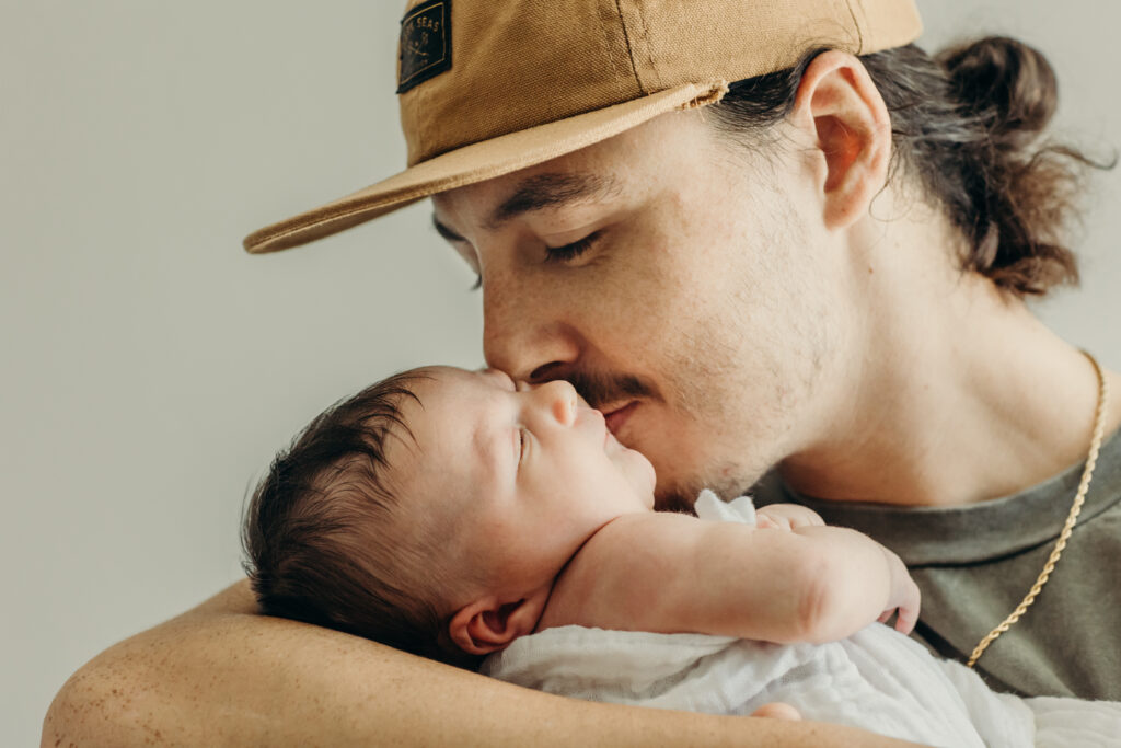 a photo from a family photoshoot of a dad wearing a yellow hat, with his hair in a bun, kissing his smiling newborn baby that is laying in his arms. 