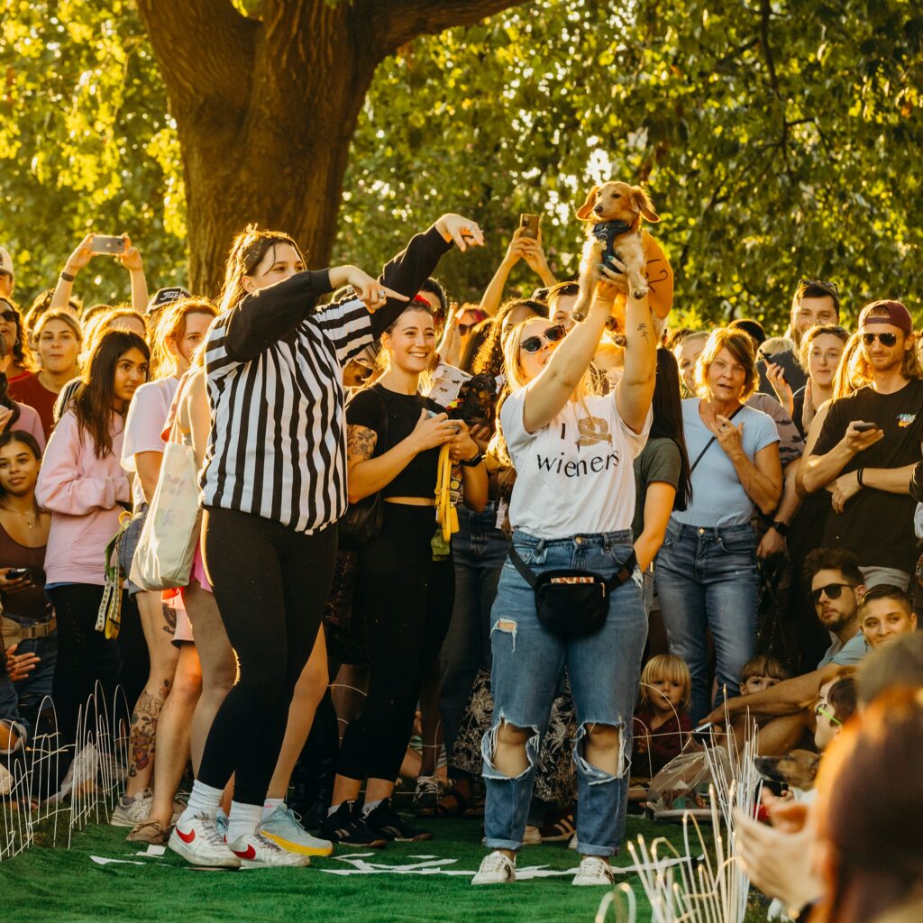 a crowd is gathered around a woman in a referee shirt and a woman in an i heart wieners shirt. the woman wearing the wieners shirt is holding her wiener dog in the air in celebration. 