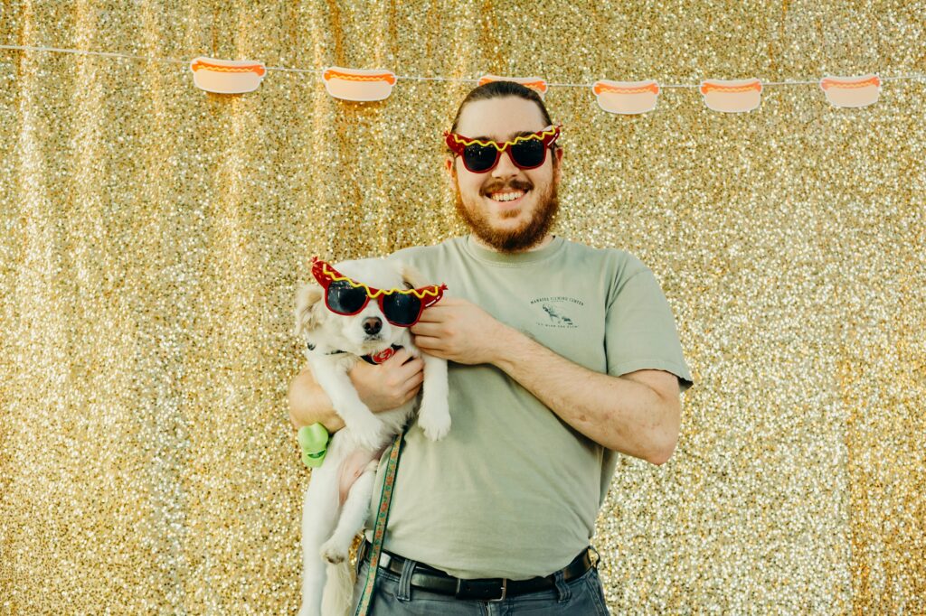 a man wearing hot dog glasses, holding his pup who is also wearing hot dog glasses. they're standing in front of a gold glittery backdrop that has hot dog cut outs in a line up top. 