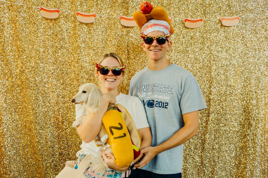 a woman wearing hot dog glasses holds her wiener dog, who is wearing a hot dog costume, stands smiling next to a man wearing a hot dog hat as well as hot dog glasses stand in front of a glittery gold background with a string of hot dog cut-outs at the top. 