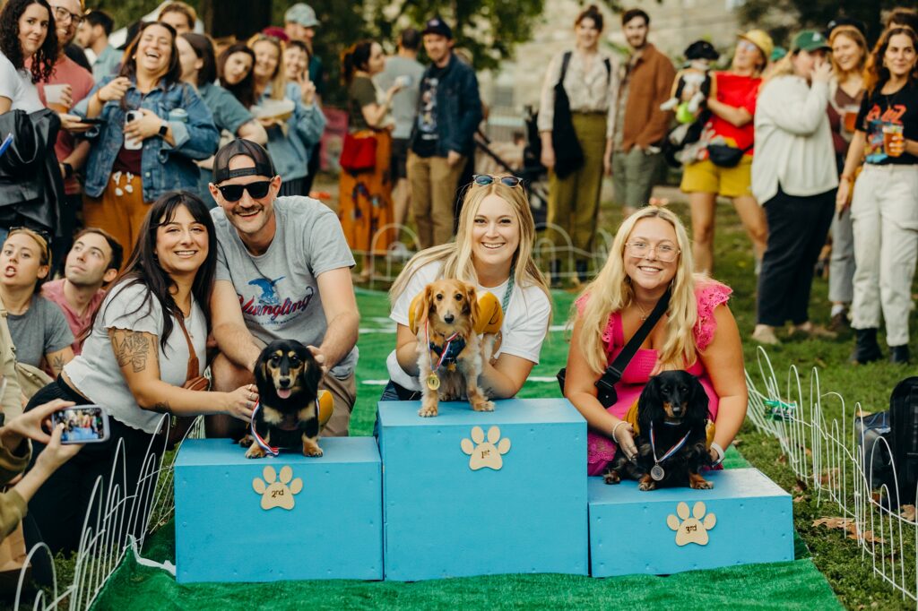 the winners of the wiener race are pictured on blue winners podiums, with pawprint signs labelling "1, 2 and 3". the winner on the 1st place podium is a tan wiener dog looking into the camera, with their blonde female owner smiling at the camera. the 2nd place winner (on the left) is a black and tan wiener dog looking into the camera, with a women with black hair and tattoos and a man wearing a backwards cap & sunglasses is behind the dog. the 3rd place winner is a black wiener dog looking into a camera as their blonde owner in a pink dress smiles at the camera.