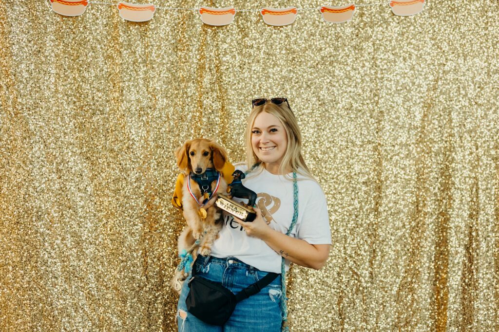 the winer race winner has a medal around their neck as they're held by his smiling blonde owner who is also holding a winer dog trophy. they are in front of a glittery gold backdrop with a string of hot dog cut outs along the top. 