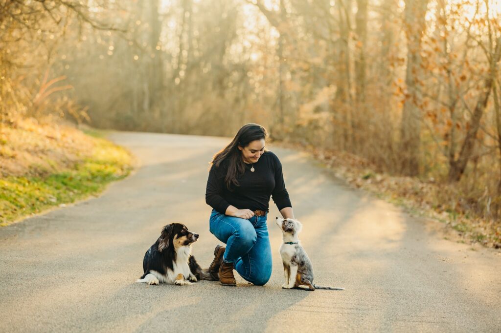 a woman kneeling on the ground, outside in the fall- smiling looking down at the puppy on her left. on her right, is her older dog looking at the two of them. 