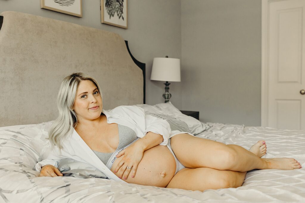 A pregnant women in a Philadelphia home sitting on her bed. She is dressed in a calvin klein bra and a white button down. 