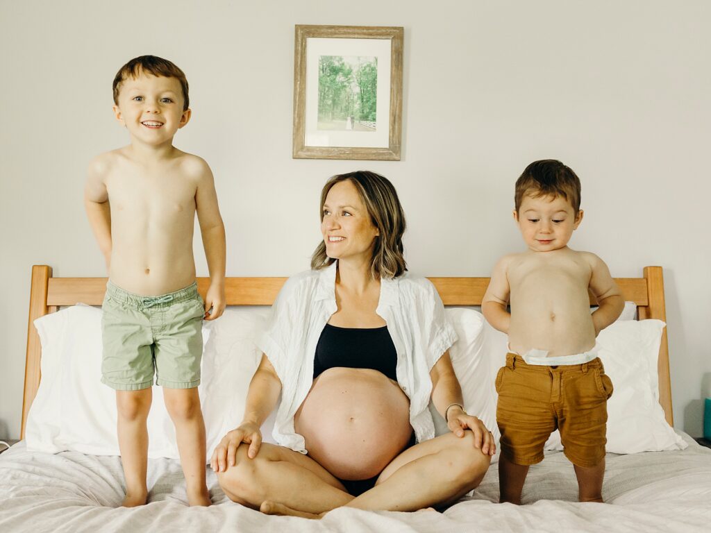 a pregnant mother sitting on her bed in a sports bra and a white button down. She is smiling to the left looking at her oldest son, while her younger son is on the right. 