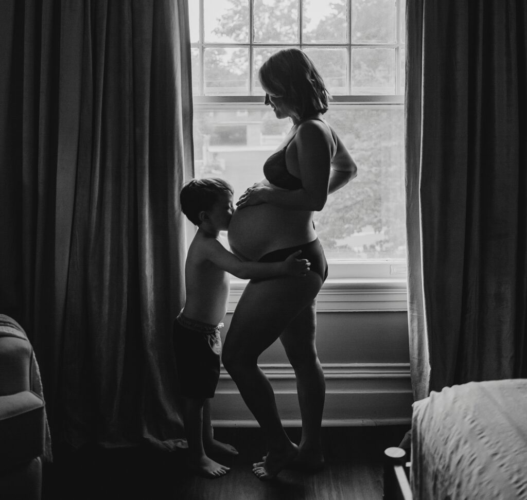 a black and white photo taken in Ambler, PA of a pregnant mother. The mother is in lingerie as her eldest son kisses her pregnant belly. 