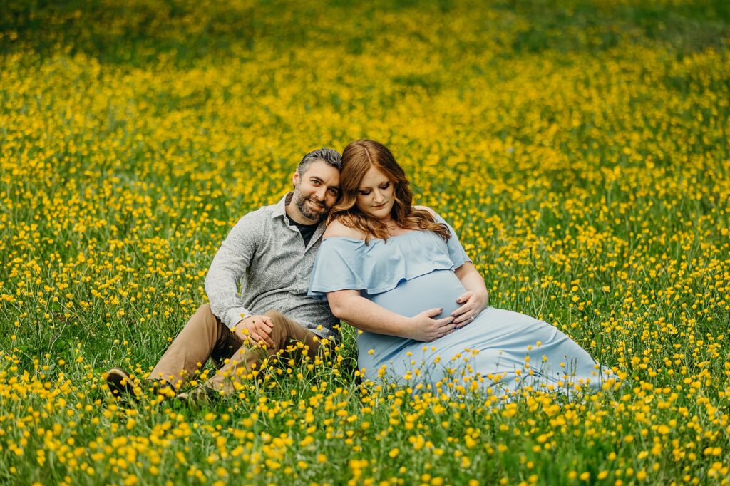 A couple sitting in a field of yellow butter cup flowers in Mt. Airy, PA. The woman is in a blue dress as she lovingly looks at her pregnant belly.