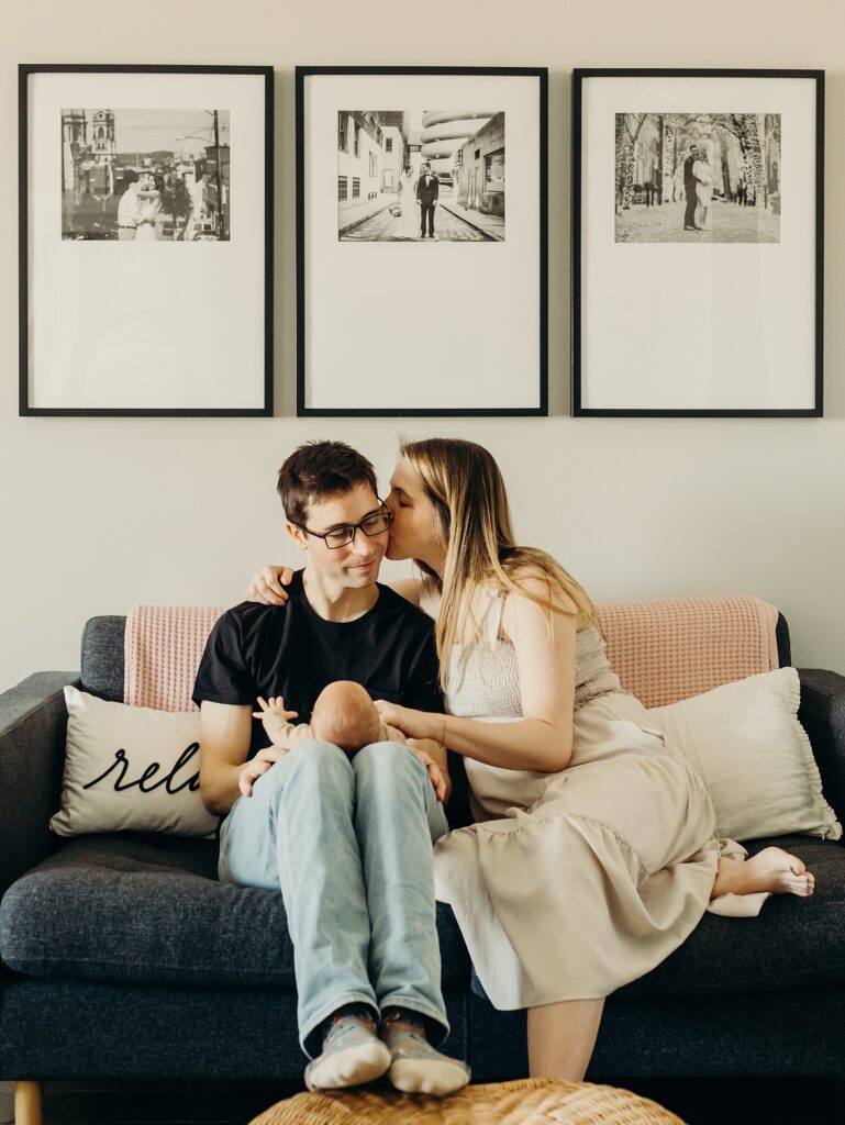 a photo of a husband and wife sitting on their dark blue couch. the wife is kissing her husbands cheek, while he smiles and looks down towards their newborn baby. on the wall behind them are 3 black and white photos in skinny black frames. the first is from their engagement , the second is from their wedding and the third is from their maternity photoshoot. 