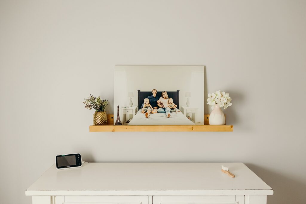 a wooden shelf is on a white wall above a white dresser. on the shelf (left to right) is a small metallic pineapple vase with flowers popping out the top, a small eiffel tower figurine, an 11x14 print of a family sitting on a bed with their 3 daughters, and a pink and white vase with white flowers. 