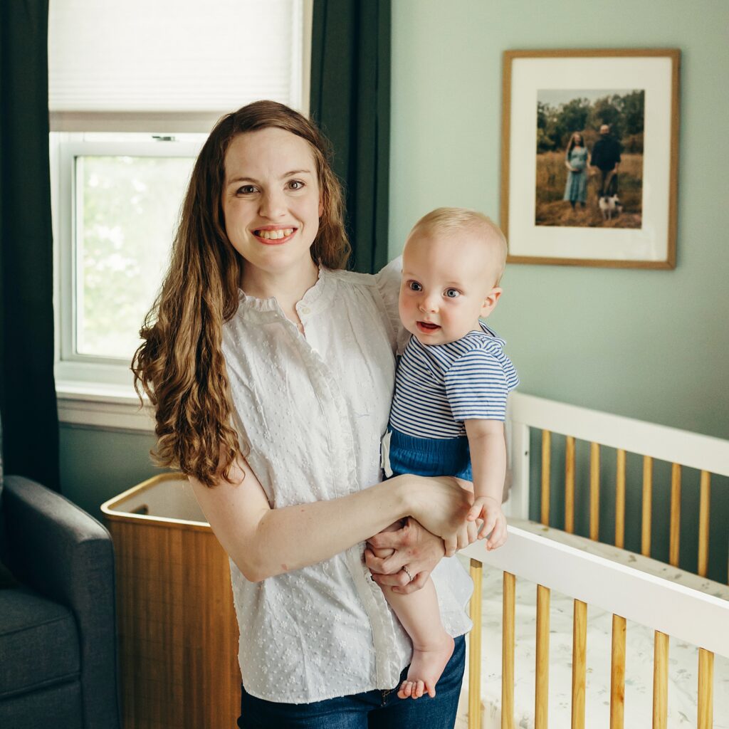 a mother is smiling, looking into the camera with her son on her hip. they are standing in the babys room, in front of his crib and a framed photo of his mother, father and dog from his parents maternity photoshoot.