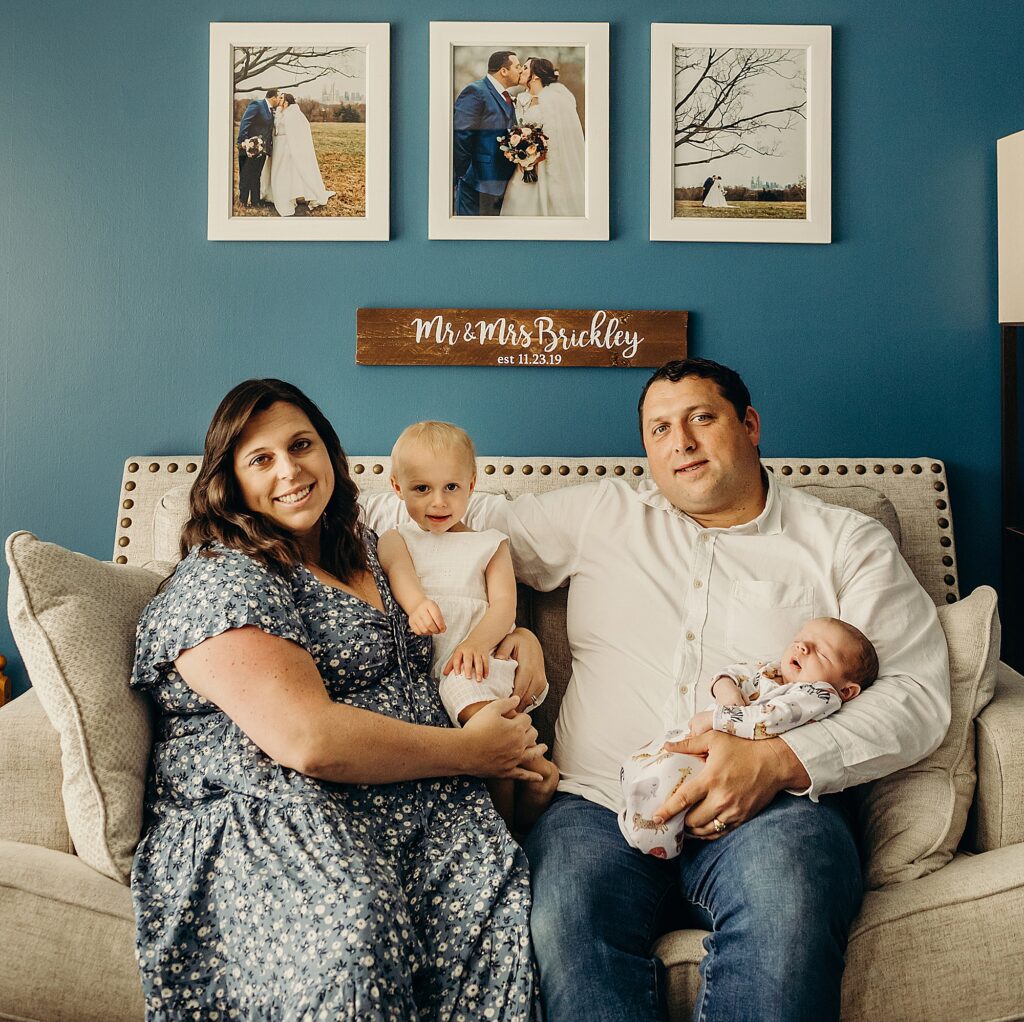 a family is sitting on their light tan couch. the mother is on the left, with her toddler daughter on her hip, while the father sits next to them holding their newborn. the newborn is asleep while the rest of the family smiles looking at the camera. on the blue wall behind them, there is 3 photos from the couples wedding framed in white frames. underneath the frames is a wooden print that says mr & mrs brickley est. 11. 23. 19