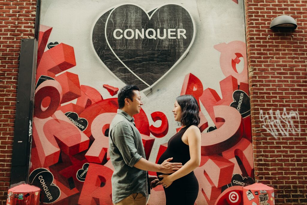 A maternity photoshoot including a couple in Fishtown, Philadelphia standing in front of the Conquer heart mural. The husband is holding his pregnant wife's belly as they smile at one another. 