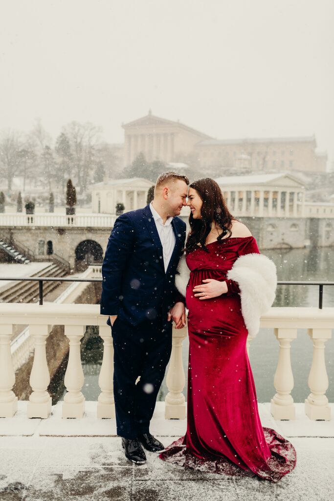 a couple smiling and posing during their maternity photoshoot while it's snowing in front of the Philadelphia Art Museum