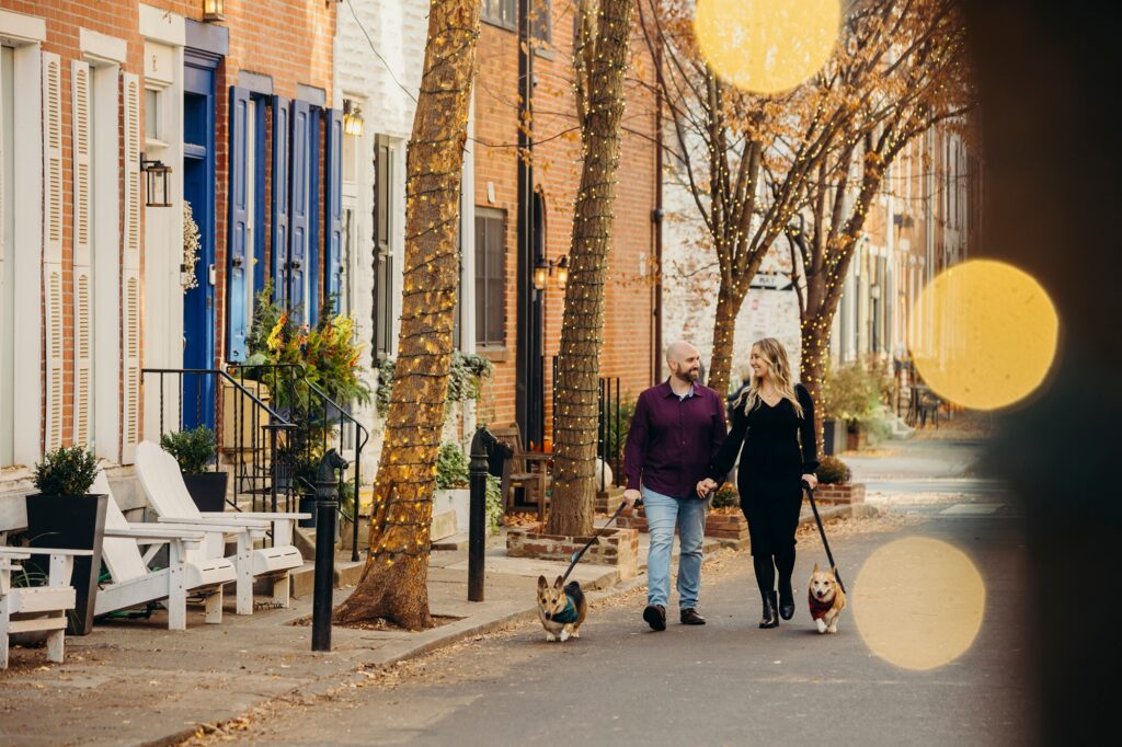 A Philadelphia couple and their two corgis walking down 17th & Addison while it's illuminated by twinkling christmas lights 