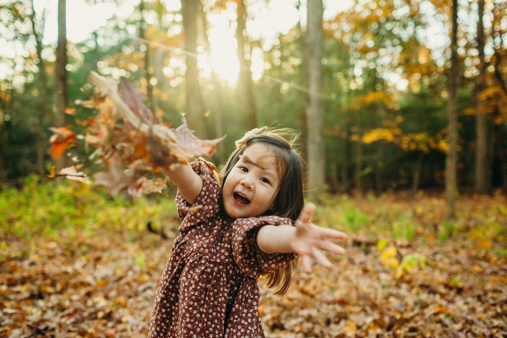 A little girl happily throwing leaves at the camera during her South Jersey fall lifestyle photo session. 